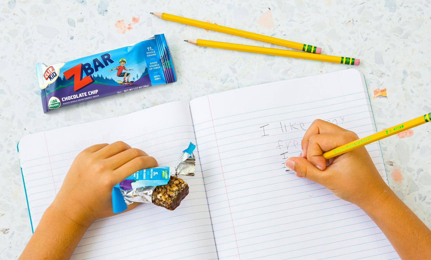Kid's hand writing in a notebook, with a CLIF Kid Zbar